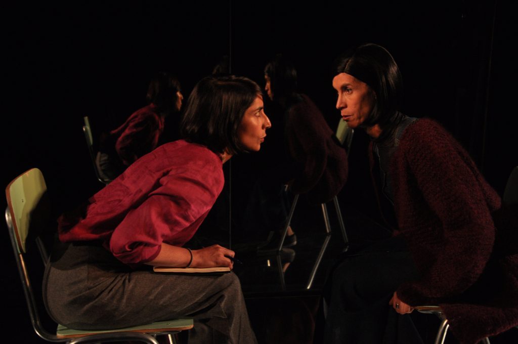 Dos actores en escena de teatro, uno sentado de perfil en silla roja, otro de pie frente a él en diálogo intenso