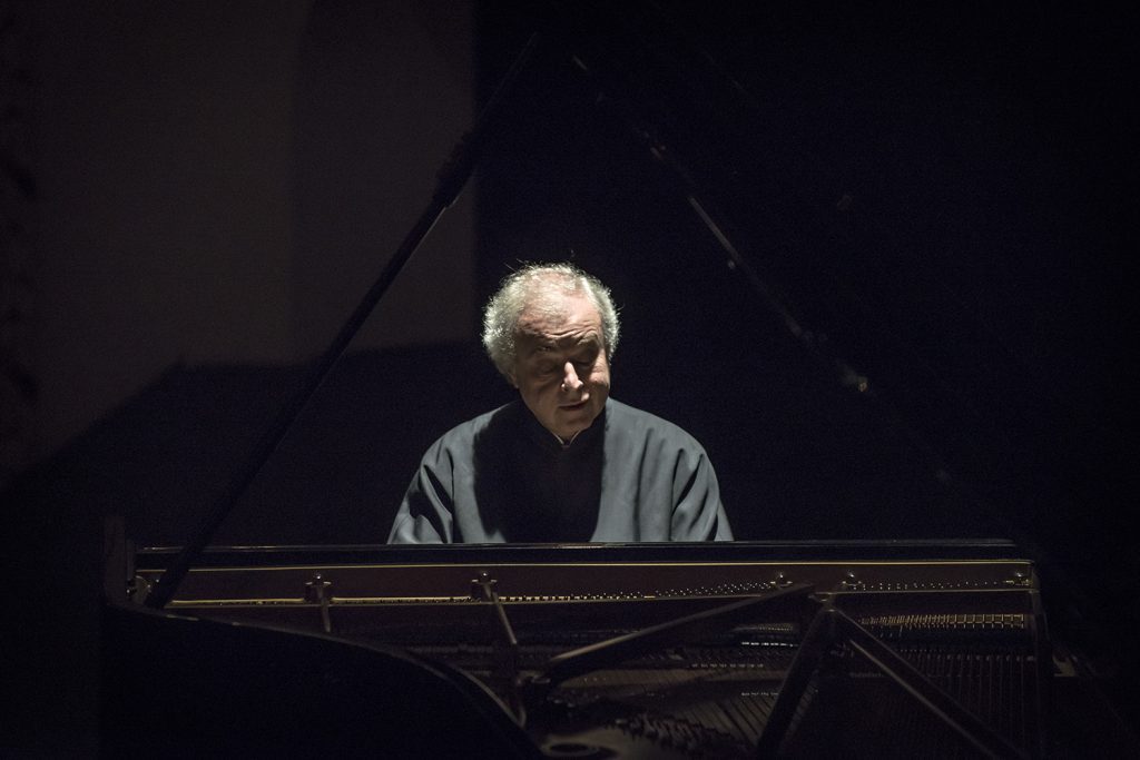 András Schiff toca el piano de cola en escenario oscuro, concentrado en la interpretación musical