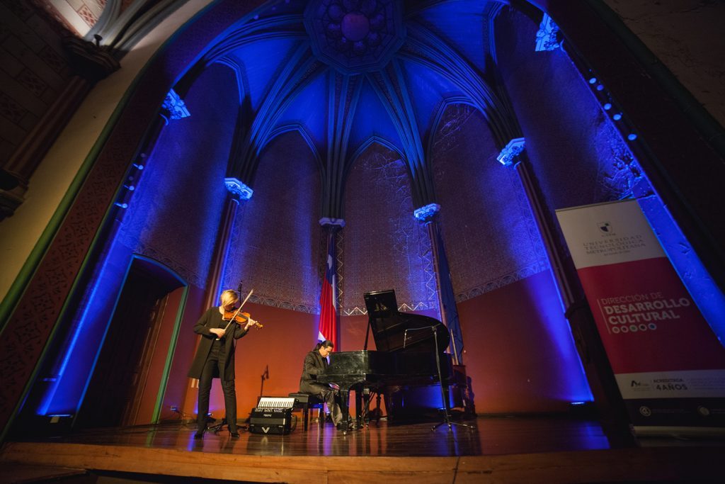 Interior de sala de conciertos con columnas azules iluminadas, soprano en escena con micrófono durante presentación