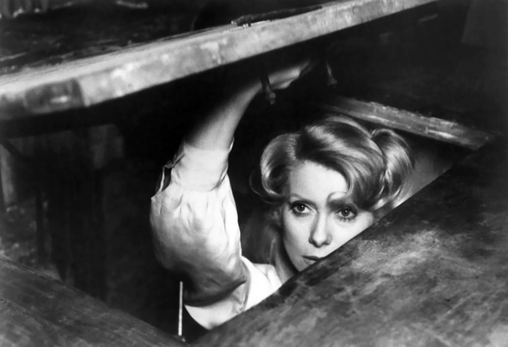 Eugenia Brito asomada bajo una mesa de madera en fotografía en blanco y negro, mirando hacia la cámara