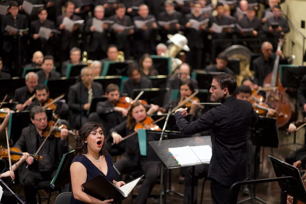Annya Pinto cantando en concierto con orquesta de fondo, dirigida por un maestro con batuta