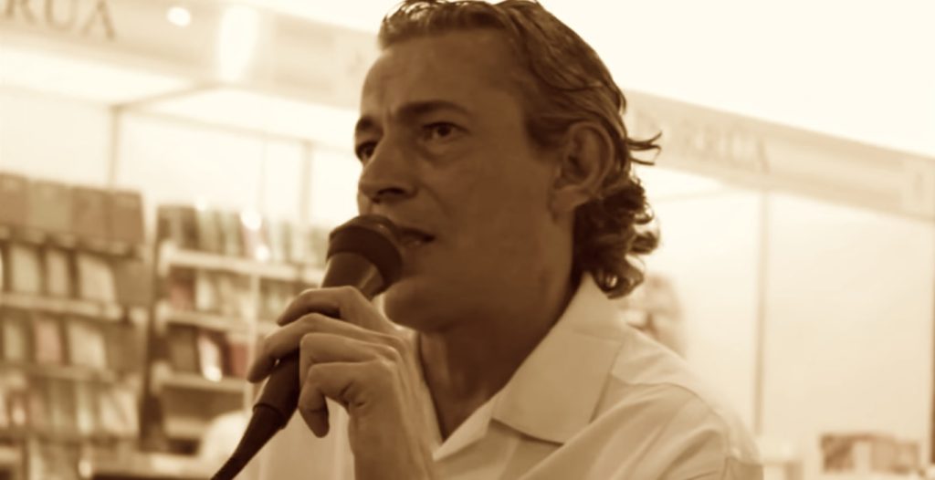 El juego de escribir: “Apuntes de un escritor malo”, por Dauno Tótoro Taulis Mauricio Bares sostiene un micrófono durante una presentación, con estanterías de libros al fondo en una biblioteca