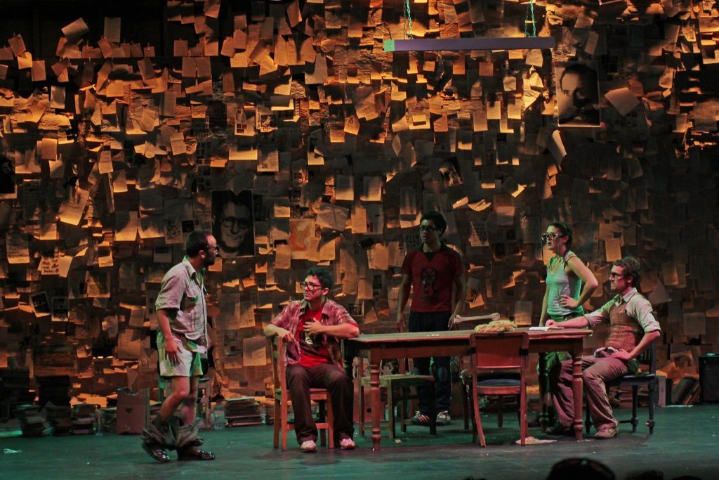 "Tratando de hacer una obra que cambie el mundo": ¿Para qué sirve hacer teatro? Cuatro actores en escena alrededor de una mesa, con pared de madera dorada de fondo, representando una obra teatral