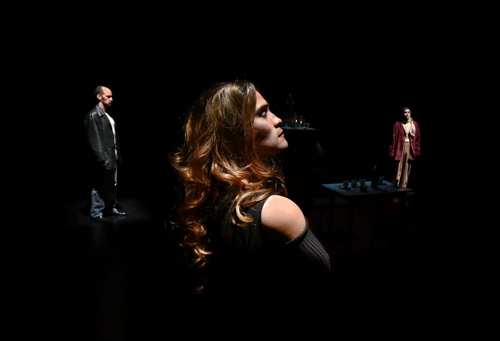 Escena teatral con mujer en primer plano de perfil y dos figuras masculinas de fondo sobre escenario oscuro