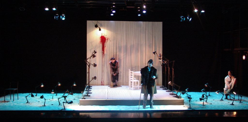 "Crimen": La dramática sutileza de la culpa Escena teatral con actor en traje oscuro en centro de escenario iluminado, acompañado por figuras secundarias