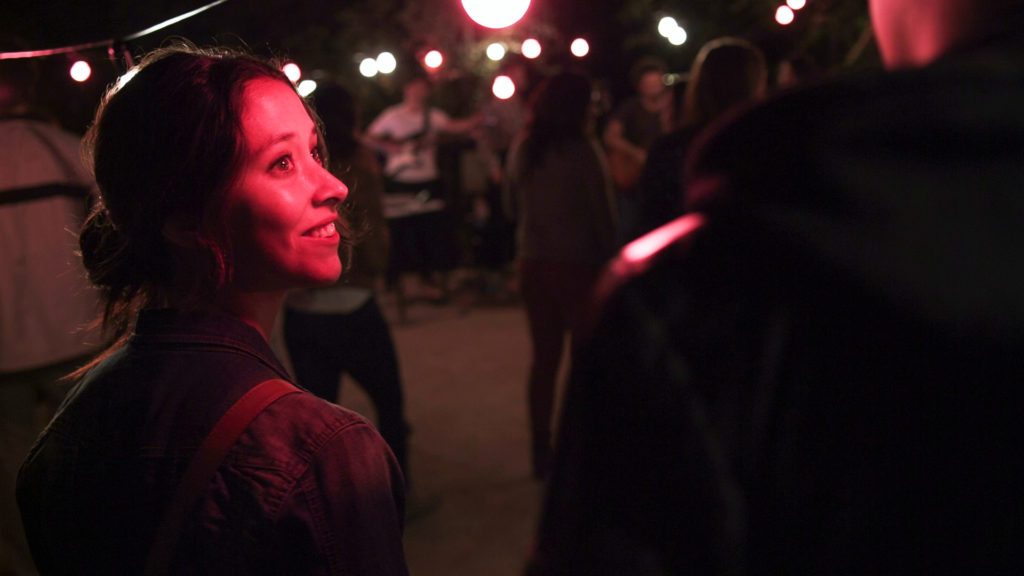 "Días de Cleo": Entre la apatía y el desajuste Mujer de perfil en ambiente nocturno con iluminación roja, rodeada de personas en lo que parece ser una escena