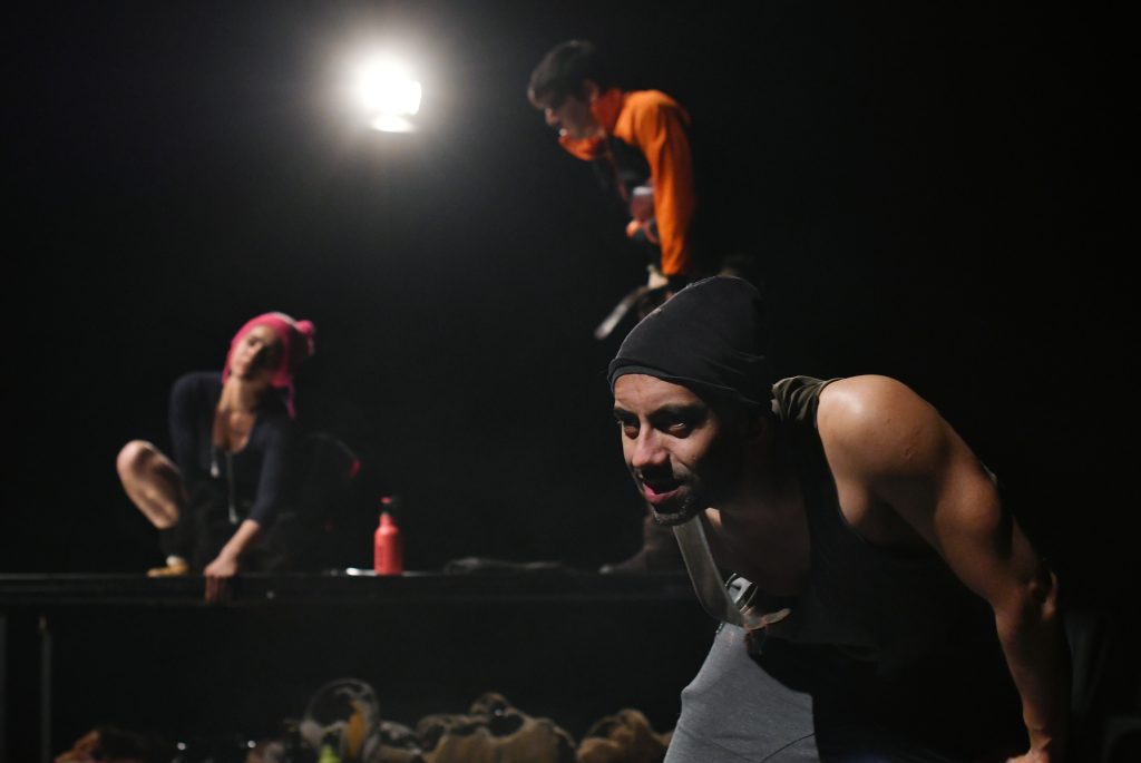 Tres actores en escena nocturna bajo luz cenital, uno de pie en posición de lunge, otros dos agachados, sobre arena