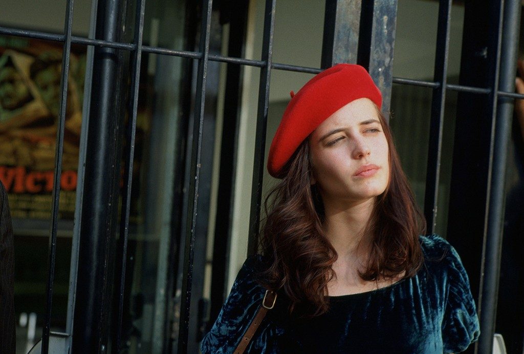 Mujer joven con boina roja y abrigo azul oscuro, mirando hacia arriba en ambiente urbano con fachada de vidrio de fondo