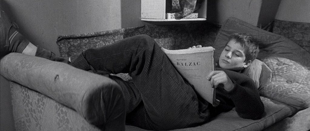 Joven acostado en sofá leyendo libro, en habitación con póster al fondo, fotografía en blanco y negro