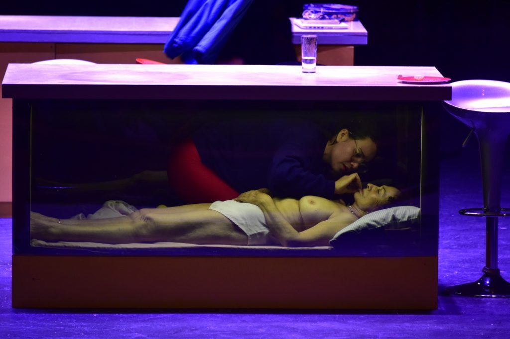 Mujer recostada en cama con neonés azul y rojo de fondo, escena íntima y doméstica iluminada