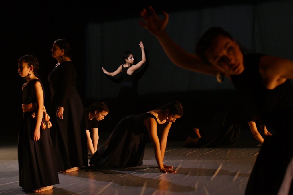 Danza "Malen" en el GAM: Un gesto de reivindicación del ser Grupo de bailarines en escena iluminados por luz cálida, ejecutando movimiento expresivo en piso oscuro durante