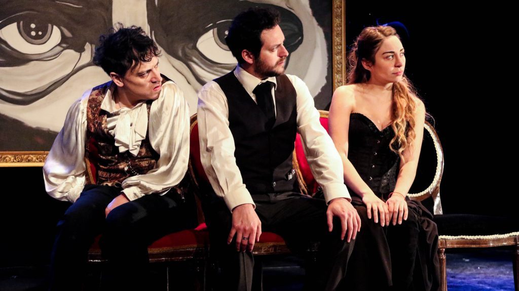 Tres actores en vestuario period en escena de teatro, sentados en sillas rojas contra fondo oscuro con decoración