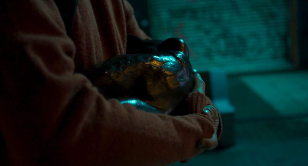 Documental "Robar a Rodin": Las paradojas del arte contemporáneo Manos sosteniendo una pequeña escultura de bronce con patina verde, iluminada contra fondo azulado oscuro