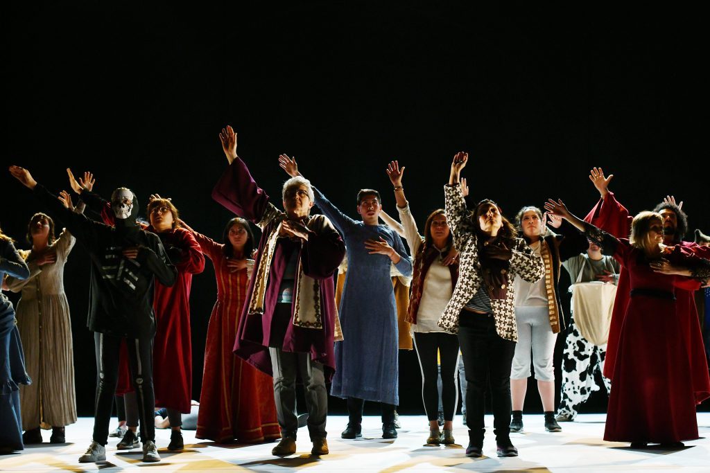 Elenco teatral en escena vistiendo trajes de época, con los brazos levantados en actitud dramática bajo iluminación