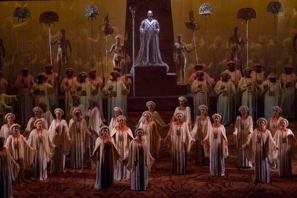 Multitud de figuras en túnicas blancas y doradas ante altar central en escena teatral de gran formato escénico