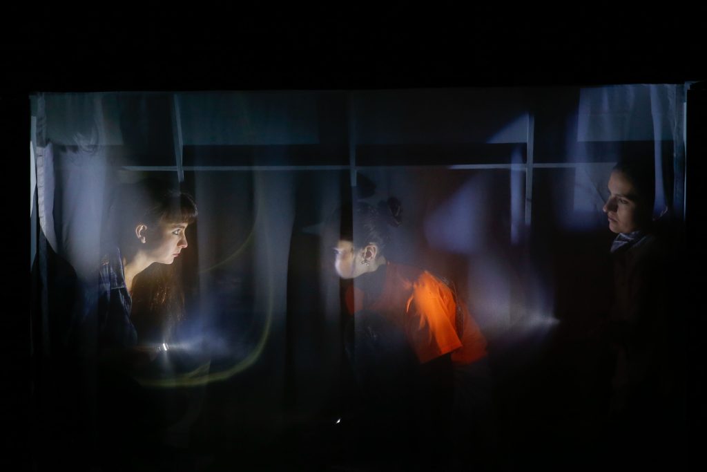 "Beben": Cuando pase el temblor Escena oscura con figuras envueltas en luz azul y naranja dentro de estructuras metálicas, ambiente de tensión
