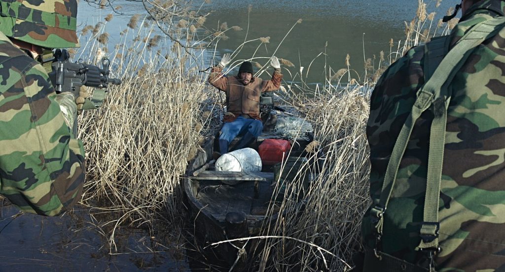“La red”: La frontera ideológica entre dos paranoias Soldados en uniforme de camuflaje persiguen a una persona entre juncos y vegetación en un entorno húmedo y pantanoso