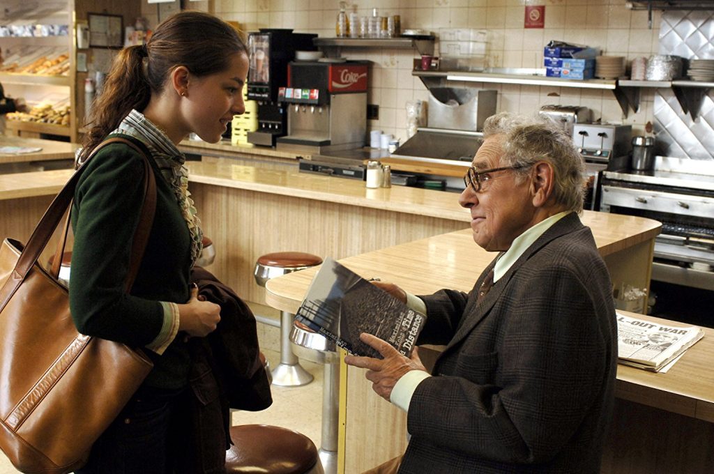 Otto Tausig conversa con una mujer en la barra de una cafetería, sostiene un libro mientras charla