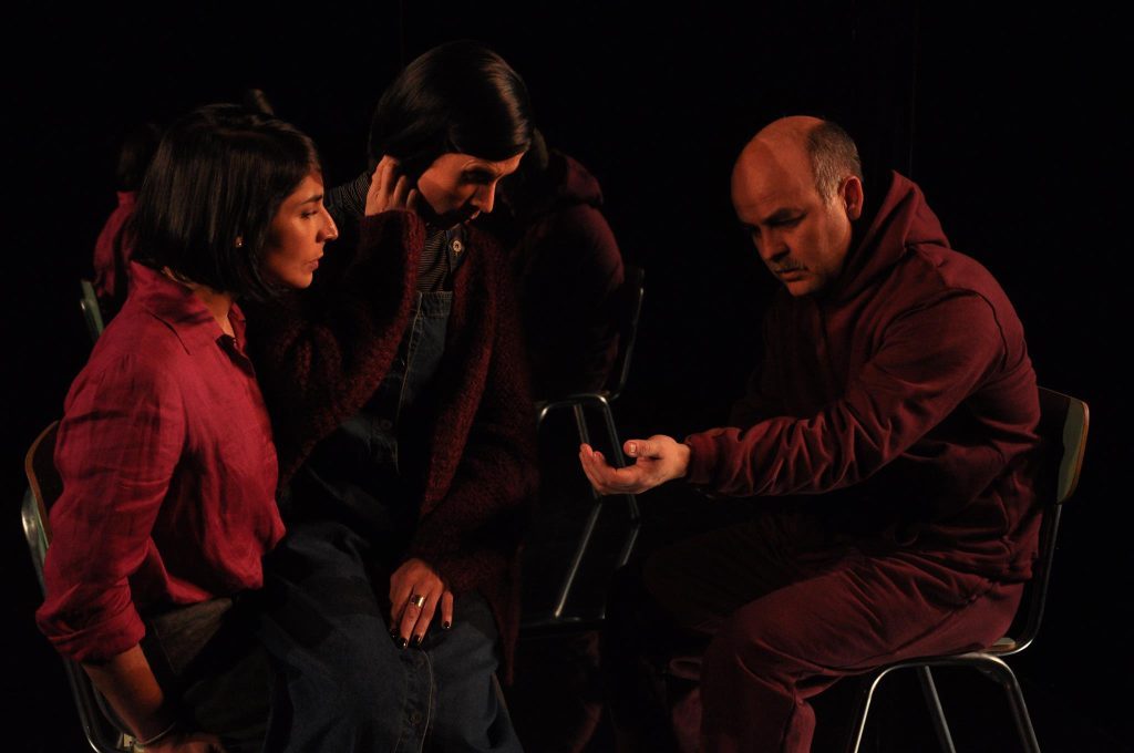 Reposición de "Prefiero que me coman los perros": El impacto emocional de un relato Tres actores en escena durante una representación teatral con iluminación roja, en momento de diálogo intenso