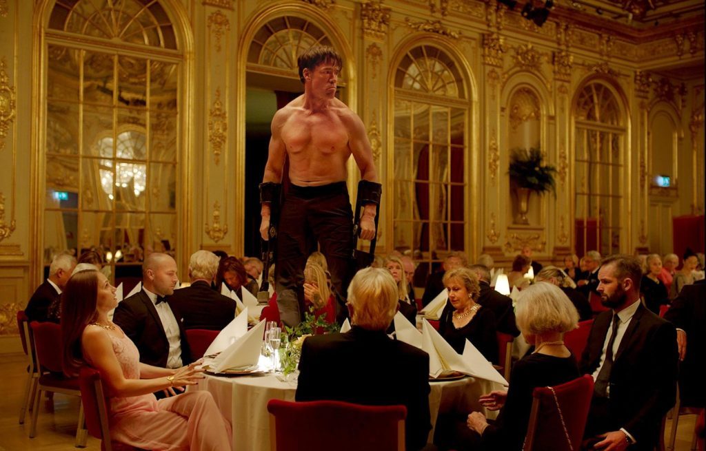 Hombre semidesnudo de pie en una sala de banquete formal mientras comensales lo observan desde sus mesas