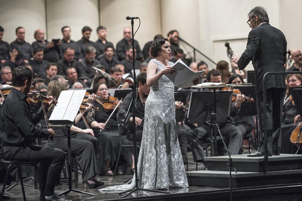 Soprano en vestido plateado canta con orquesta sinfónica y director de espaldas en concierto dedicado a Prokófiev