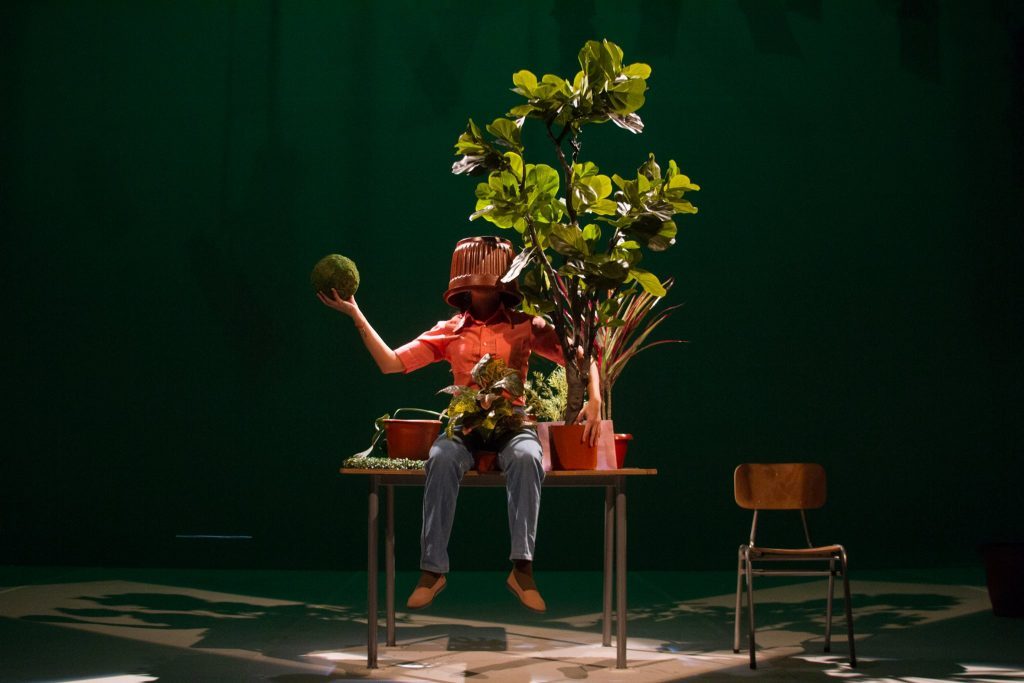 Actor sentado en silla de madera sosteniendo maceta con planta amarilla sobre fondo verde oscuro de teatro