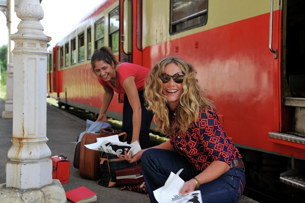 “Tiempo de revelaciones”: La eternidad de un instante Dos mujeres sentadas en escalinata de tren rojo, una sostiene documento, sonríen hacia cámara en día soleado