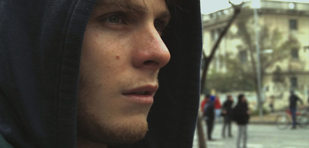Un capítulo de la novela "El vestigio del silencio", del cineasta Jorge Yacomán Primer plano de rostro de hombre con capucha oscura en entorno urbano, de perfil hacia la izquierda, con transeúntes