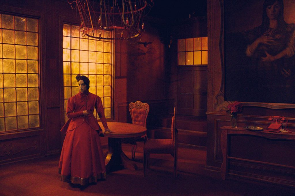 Mujer en vestido rojo de pie en interior iluminado con tonos cálidos, junto a silla de madera y ventanas con vidrios
