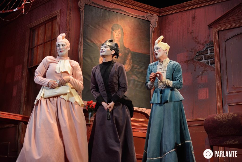 Tres actrices en escena de teatro con vestuario de época en tonos pastel y negro, sobre fondo rojo oscuro con pintura