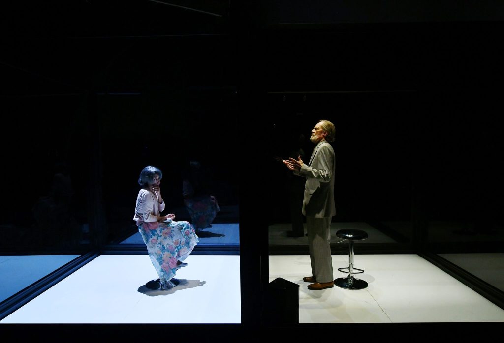Dos actores en escena iluminada: uno sentado a la izquierda, otro de pie a la derecha en posición de monólogo
