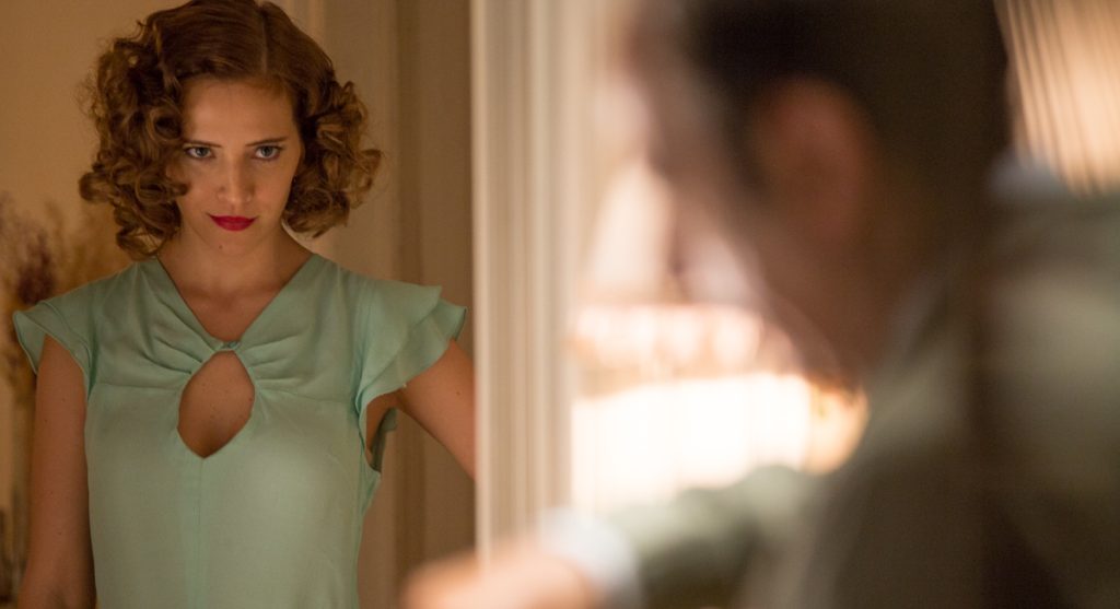 “Los que aman, odian”, un plan de evasión Mujer con cabello rizado ondulado y labios rojos, vistiendo blusa verde vintage, mirando hacia un lado en interior