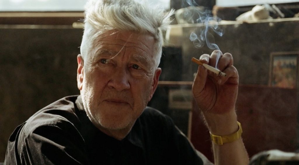 David Lynch con cigarrillo en mano, gesto pensativo, interior con objetos artísticos de fondo