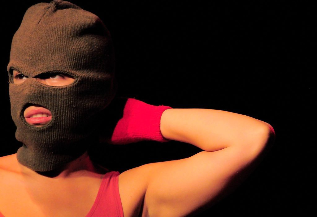 Persona con pasamontañas negro y top rojo posando contra fondo oscuro, expresión dramática y teatral