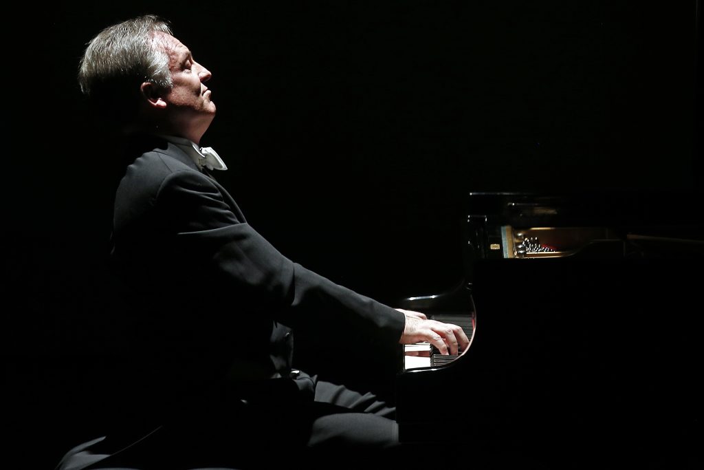 Alfredo Perl, pianista de perfil, tocando el piano en escena iluminada contra fondo negro