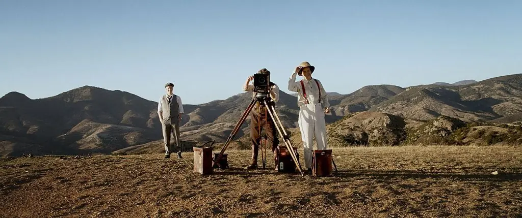Equipo de producción cinematográfica montando cámara en trípode en paisaje árido desértico con montañas al fondo