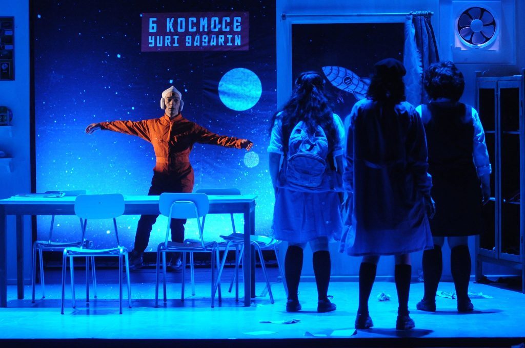 Escena de teatro con actriz en plataforma elevada bajo iluminación azul, rodeada de espectadores en penumbra, ambiente