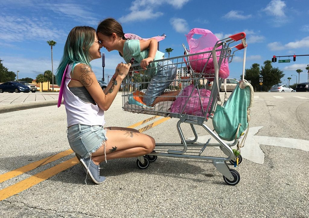 “Proyecto Florida”, el resplandor inalcanzable del oeste Mujer arrodillada junto a carrito de compras lleno de objetos coloridos en estacionamiento bajo cielo despejado