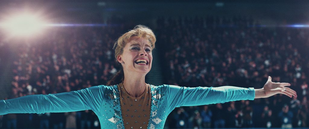 "Yo, Tonya": La violencia de las carencias Tonya Harding con los brazos extendidos en traje de patinaje azul turquesa bajo focos de arena de hielo