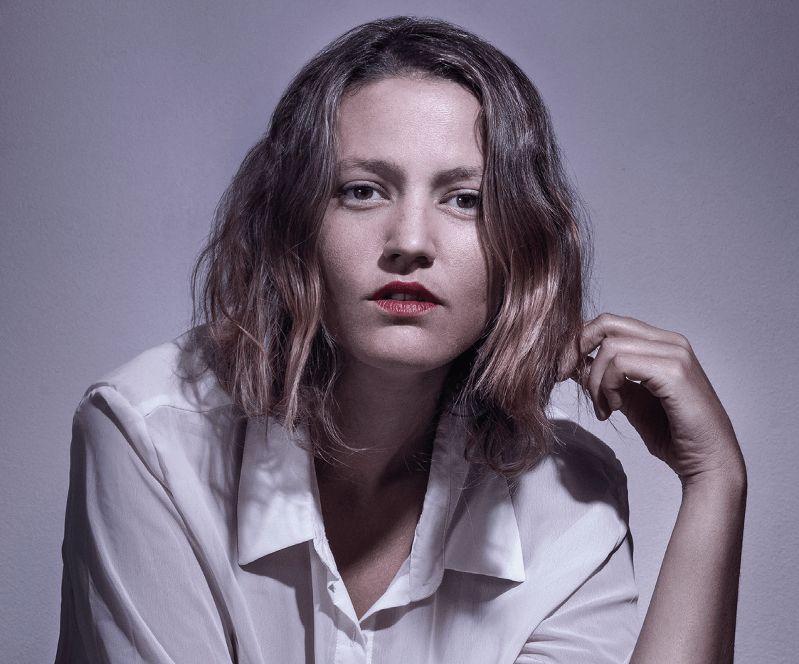 "El final del sendero", de Carolina Brown: La vida en movimiento Retrato de Carolina Brown, escritora, con cabello castaño ondulado y blusa blanca sobre fondo gris neutro