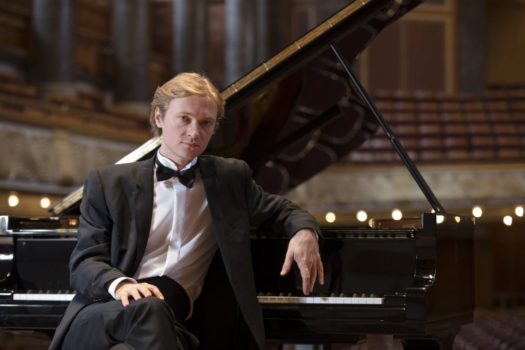 Pianista Dmitri Levkovi posando junto al piano de cola en sala de conciertos con decoración de luces doradas