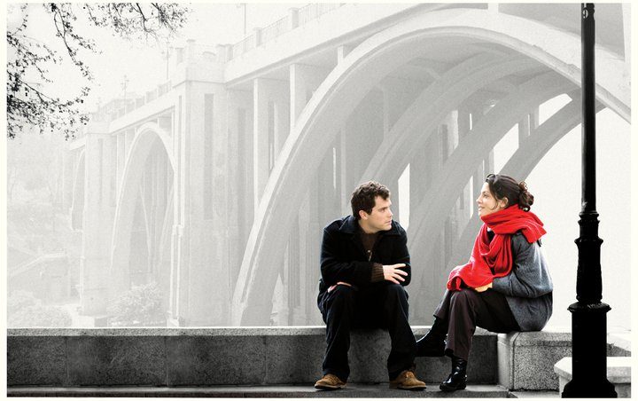 Jonás Trueba en conversación con una mujer de chaqueta roja, sentados bajo un arco de arquitectura moderna