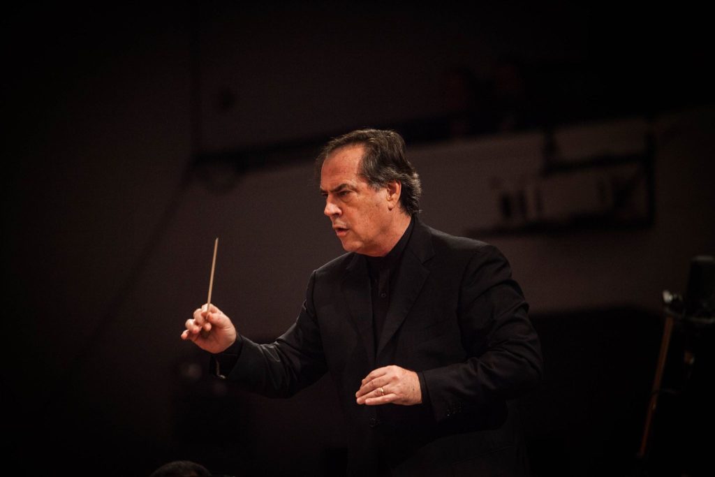 Maximiano Valdés vuelve a dirigir a la Orquesta Sinfónica Nacional de la mano de violinista amante del rock Maximiano Valdés dirigiendo con batuta, vistiendo traje negro, en escenario de concierto con iluminación oscura