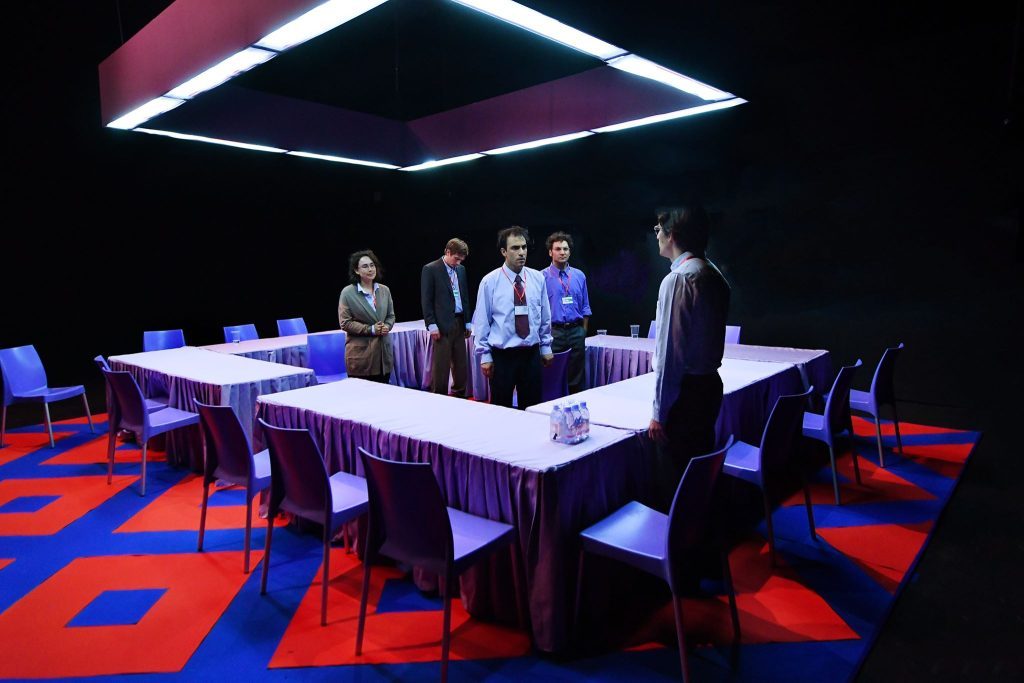 Escena de teatro con cinco actores en un escenario minimalista, piso azul y rojo, bajo iluminación neón púrpura