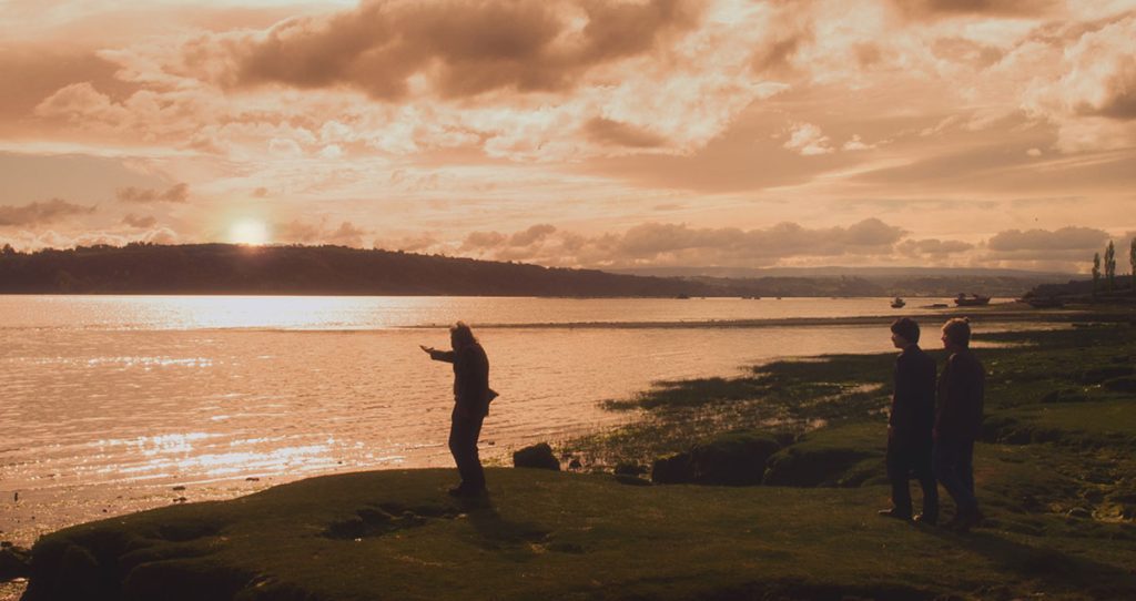 "...Y de pronto el amanecer": La fotografía de un Chile inexistente y periférico Hombre en la orilla de un lago al atardecer, con cielo dorado y reflejo en el agua, paisaje rural chileno