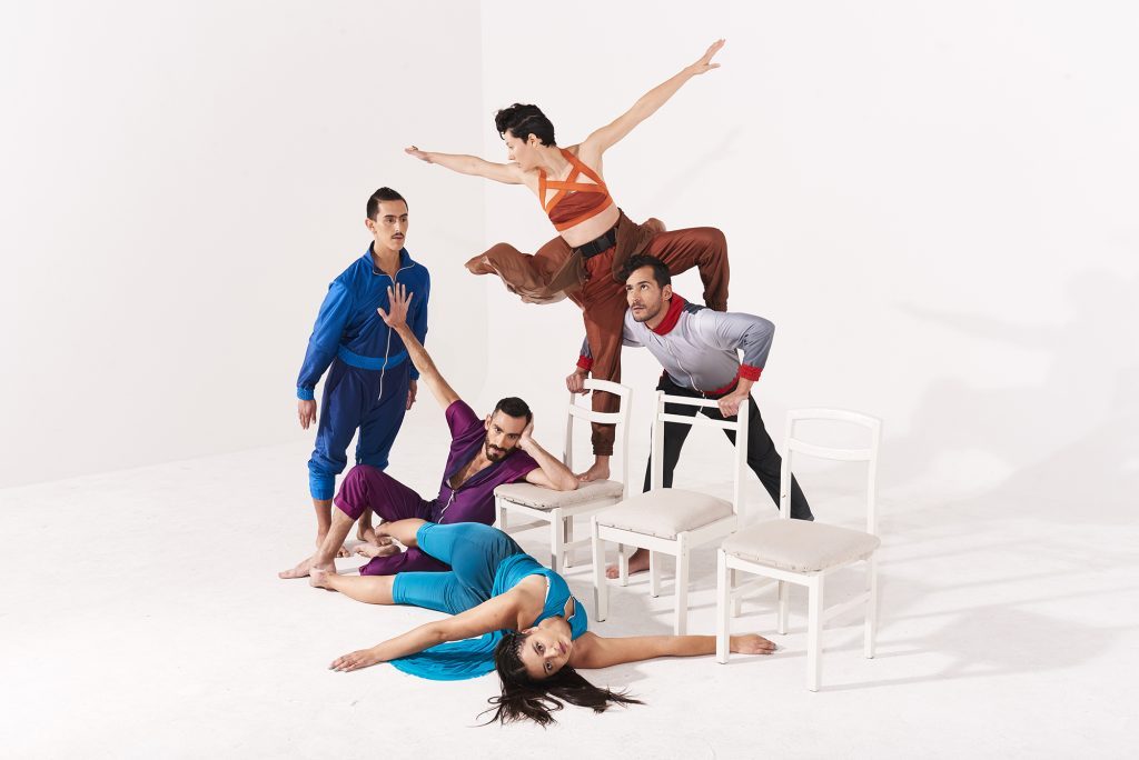 Grupo de performers en movimientos corporales dinámicos sobre plataforma blanca, vistiendo ropa de colores, en escena