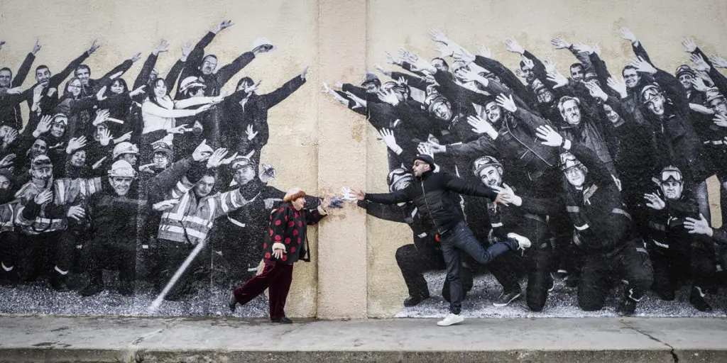 Dos figuras en blanco y negro frente a mural con siluetas de personas levantando los brazos, separadas por pared