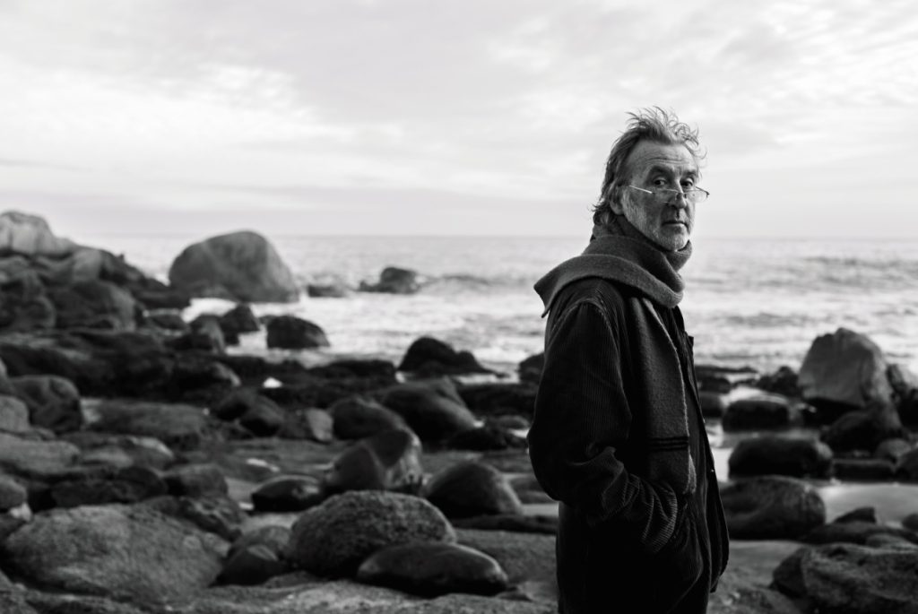 Claudio Bertoni, poeta chileno, de perfil junto a rocas en playa con mar de fondo en fotografía blanca y negra