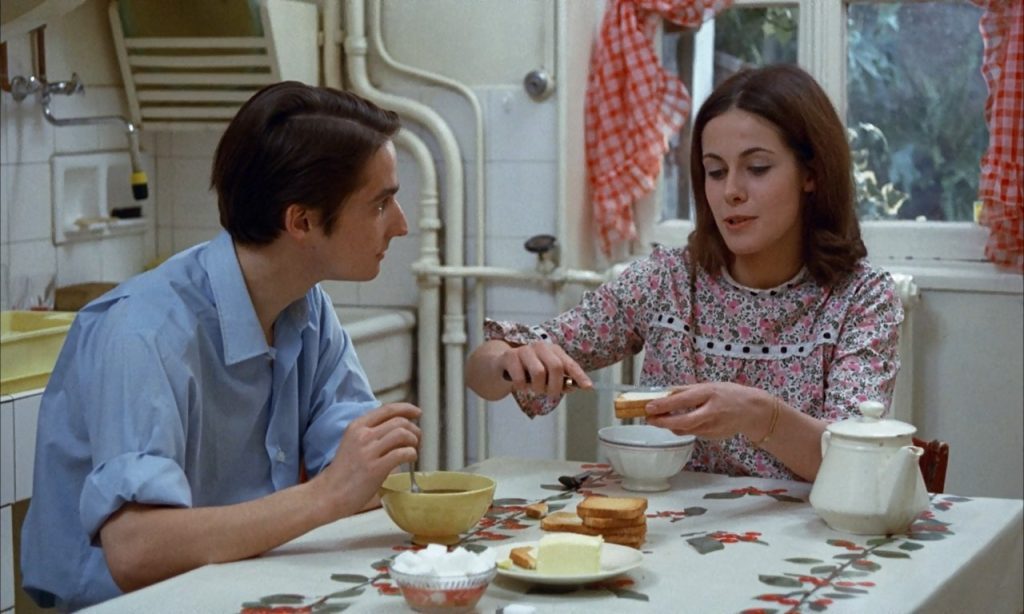 Dos personas en una cocina comparten una comida; él en camisa azul y ella sirviendo comida en un plato, ambiente