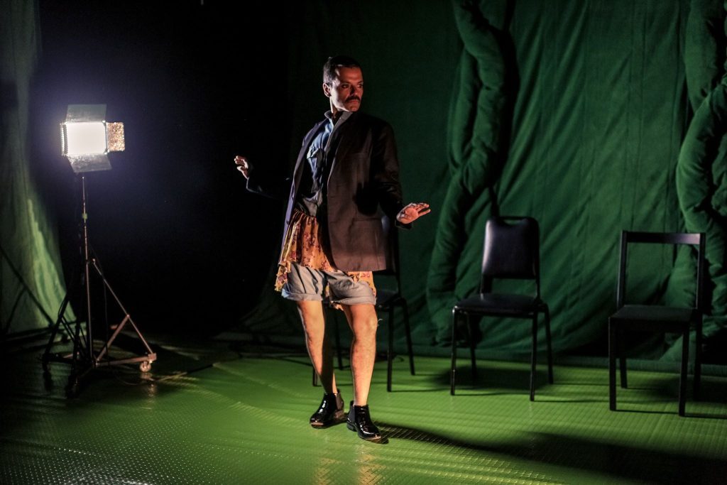 Actor en escena de teatro bajo luz frontal, vistiendo chaqueta marrón y pantalones cortos, sobre piso verde con set